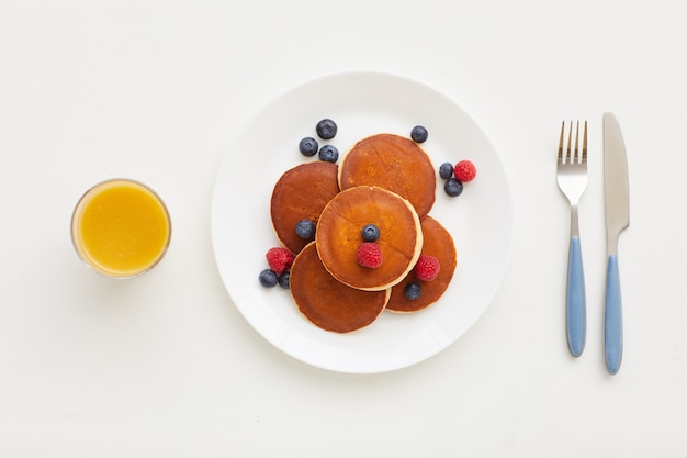 Vista dall'alto alla composizione minima di deliziose frittelle dorate con frutti di bosco freschi accanto a succo d'arancia e coltello e forchetta, concetto di colazione