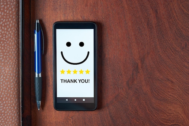 Photo top view of mobile phone with smiling happy face and five stars rating with the text thank you on white screen pen and notebook lying on dark brown wooden table concept of positive feedback