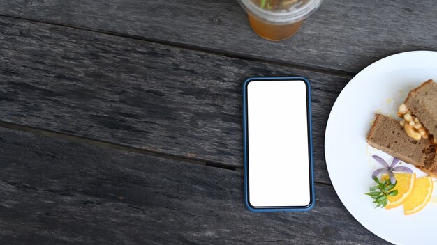 Top view of mock up smart phone with blank screen and cake on wooden table at cafe.