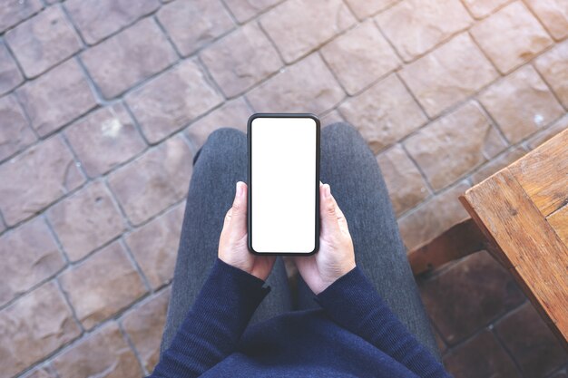 Top view mockup image of hands holding black mobile phone with blank white screen while sitting in the outdoors