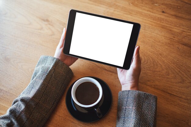 Top view mockup image of hands holding black tablet pc with blank white screen with coffee cup on wooden table