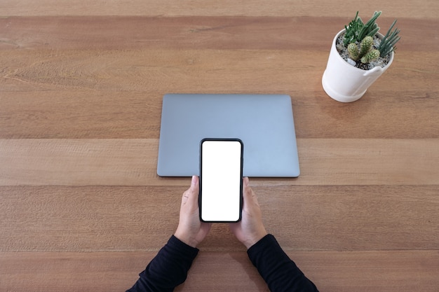 Top view mockup image of a hands holding a blank white screen mobile phone and laptop on wooden table in office