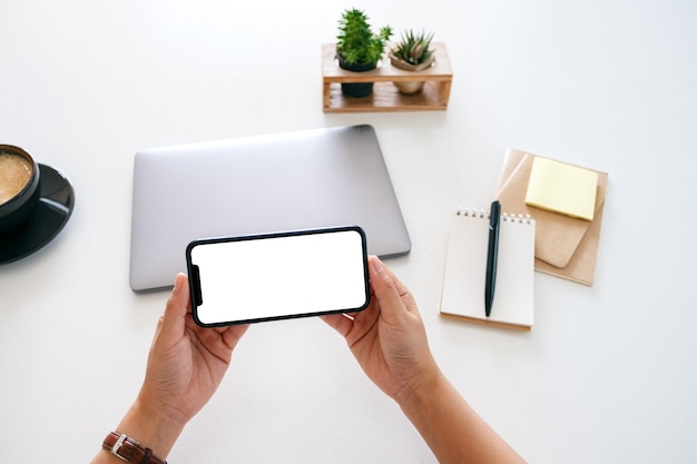 Immagine mockup vista dall'alto delle mani che tengono il telefono cellulare con schermo vuoto e computer portatile sul tavolo in ufficio