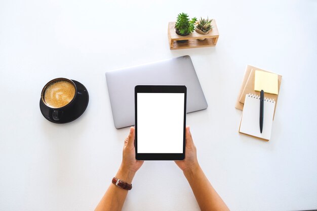 Top view mockup image of hands holding a tablet with blank white screen and laptop computer on the table in office