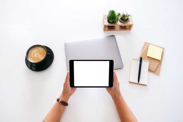 사무실의 나무 테이블에 빈 흰색 화면과 노트북 컴퓨터가 있는 태블릿을 들고 있는 손의 상위 뷰 모형 이미지
