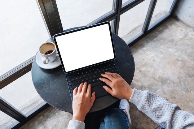 Top view mockup image of hands using and typing on tablet keyboard with blank white desktop screen as computer pc with coffee cup on the table