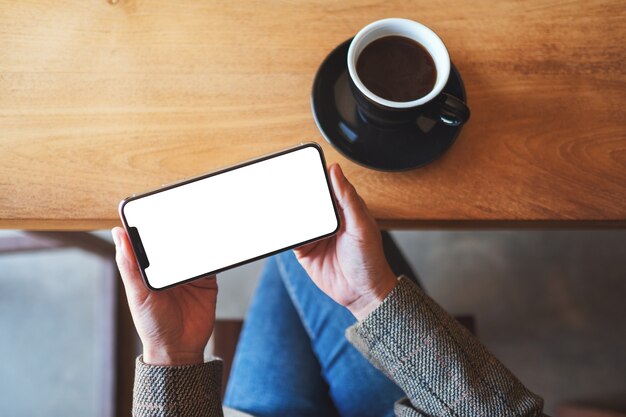 Top view mockup image of a woman holding black mobile phone with blank desktop screen while sitting in cafe