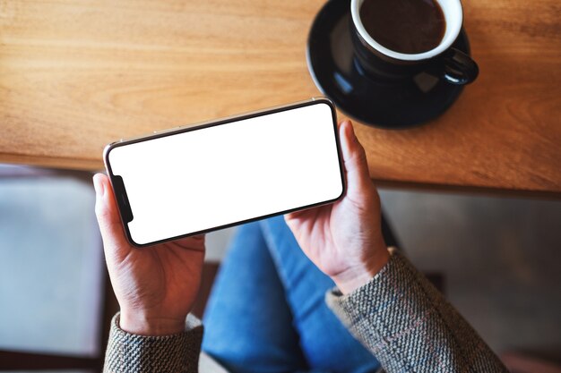 Top view mockup image of a woman holding black mobile phone with blank desktop screen while sitting in cafe