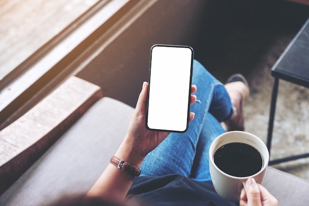 座ってカフェでコーヒーを飲みながら空白の画面で黒い携帯電話を保持している女性の上面モックアップ画像