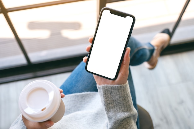 커피 컵이 있는 빈 흰색 데스크탑 화면이 있는 검은색 휴대폰을 들고 있는 여성의 상위 뷰 모형 이미지