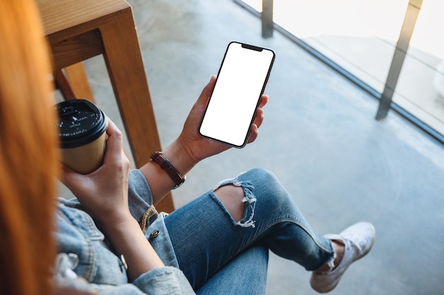 Top view mockup image of a woman holding a black mobile phone with blank white desktop screen with coffee cup