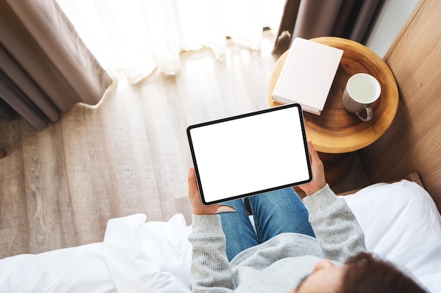 自宅で居心地の良い白いベッドに座っている間、空白のデスクトップの白い画面で黒いタブレットPCを保持している女性の上面モックアップ画像