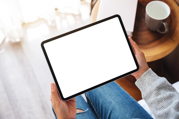 Top view mockup image of a woman holding black tablet pc with blank desktop white screen while sitting on a cozy white bed at home