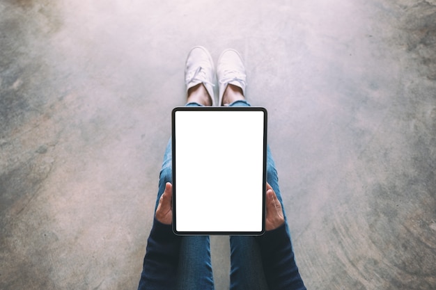 Foto immagine mockup vista dall'alto di una donna che tiene in mano un tablet pc nero con schermo bianco vuoto mentre è seduta sul pavimento