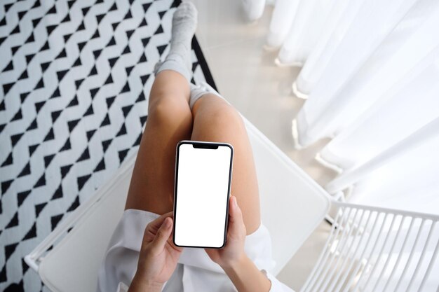 リラックスした気分で寝室に座っている間空白のデスクトップの白い画面で携帯電話を保持している女性の上面モックアップ画像