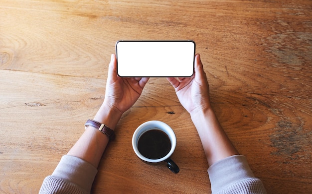 Top view mockup image of a woman holding mobile phone with blank white desktop screen with coffee cup on wooden table