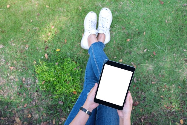 Top view mockup image of a woman holding and using black tablet pc with blank white desktop screen while sitting in the outdoors