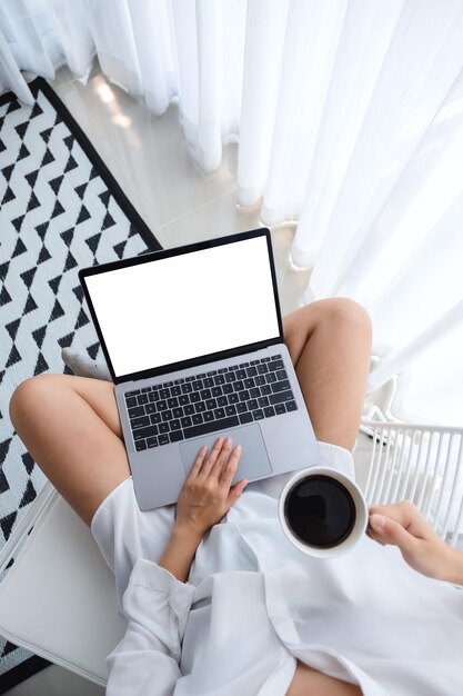 Top view mockup image of a woman using laptop computer with blank screen while sitting and drinking coffee in a bedroom at home