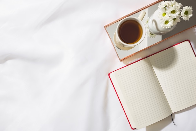 Top view of morning scene in voile fabric background with a red notebook, mug of tea and a vase of white flowers in a mirrored brass tray, with space for text