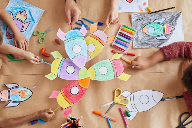 Foto vista dall'alto di un gruppo multietnico di bambini in possesso di immagini di razzi spaziali mentre si godono lezioni di arte e artigianato in età prescolare o centro di sviluppo