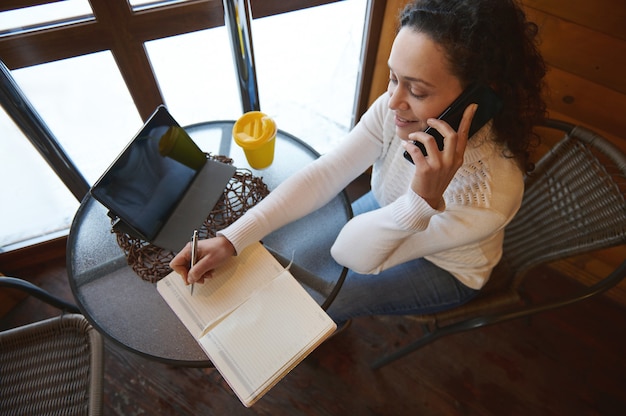 Вид сверху на переговаривающуюся женщину по телефону и запись в дневнике, сидя у окна в деревянном кафе
