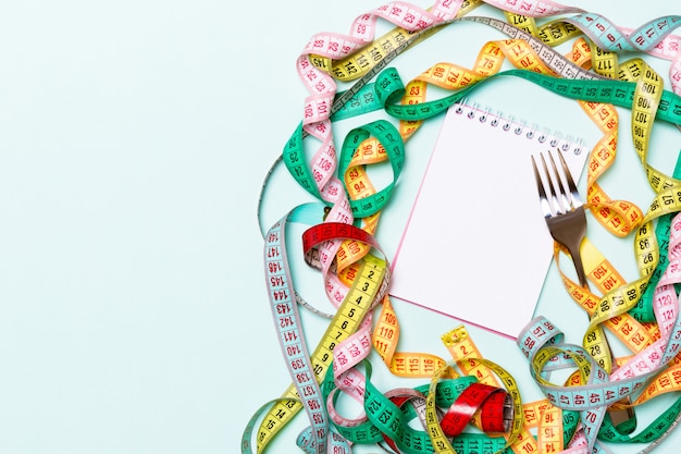 Top view of notebook with fork surrounded with colorful measuring tapes on blue background