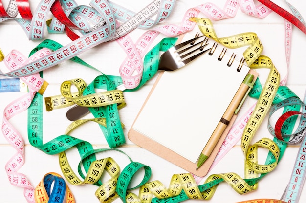 Top view of notebook with fork surrounded with colorful measuring tapes on white wooden . copyspace with diet planning