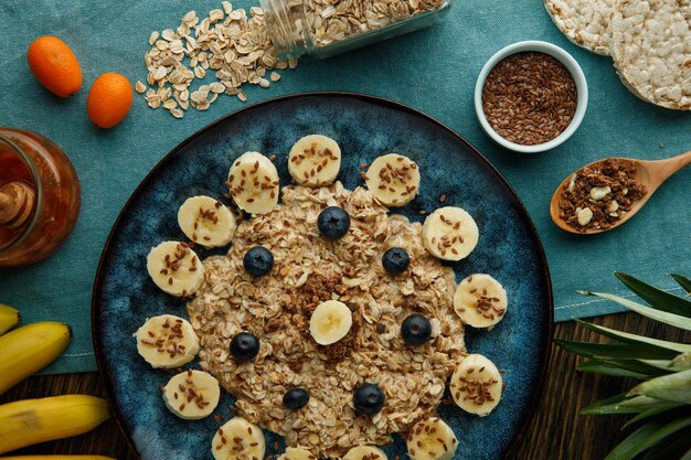 Photo top view of oatmeal with banana blackthorn walnut and sesame in plate with jam oat kumquat pineapple leaves crispbread on blue cloth on wooden background