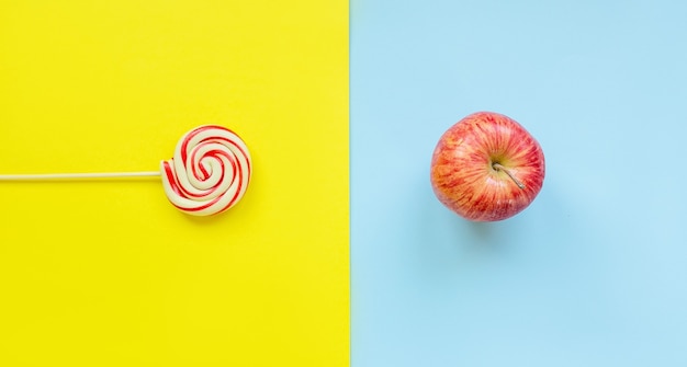 Фото Вид сверху конфеты на палочке против свежего красного яблока на ярком двухцветном ярком желто-синем фоне. плоская диета и концепция здорового образа жизни