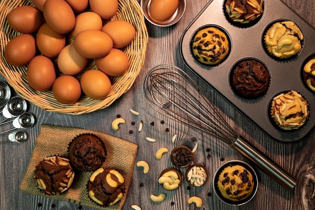 新鮮な自家製のおいしいバニラとチョコレートマフィンの紙カップケーキホルダー材料卵耐熱皿パンアーモンドスライスchooclateチップと木製の背景に分離されたカシューナッツの上面図