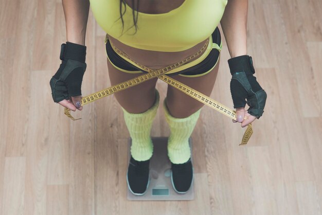 Фото Вид сверху довольно стройной женщины с мерками вокруг тела, стоящей на весах. похудение в тренажерном зале. фитнес-женщина проверяет результаты после тренировки в оздоровительном клубе. спортивное взвешивание.