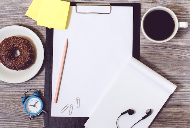 Vista dall'alto di accessori per ufficio su scrivania in legno: orologio, tazza di caffè, appunti, ciambella, blocco note e adesivi per colla