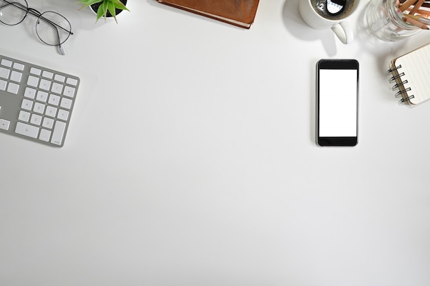 Top view office desk mockup smartphone, computer keyboard, coffee, notepad paper on white table.