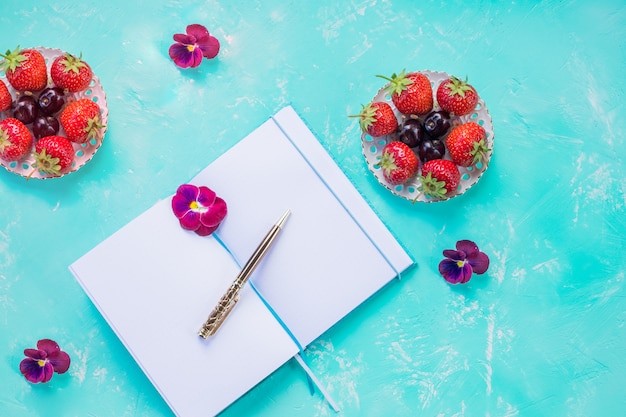 열린 빈 노트북의 상위 뷰는 파란색 책상 벽 위에 조롱. 와일드 베리 과일 배열. 딸기, 바쁜 아침 식사의 개념, 여름 플래너를 할 목록.