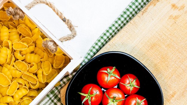 Top view of pasta in a wooden crate and fresh ripe cherry tomatoes in a black bowl on a rustic white wooden table ingredients and food concept