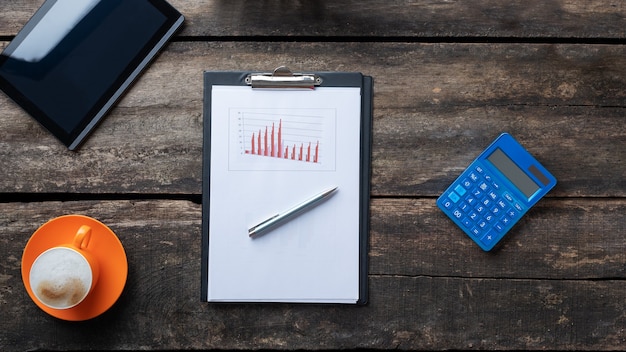 Top view of pen lying on a paperwork with graphs and charts on a desk next to digital tablet, calculator and cup of fresh coffee.