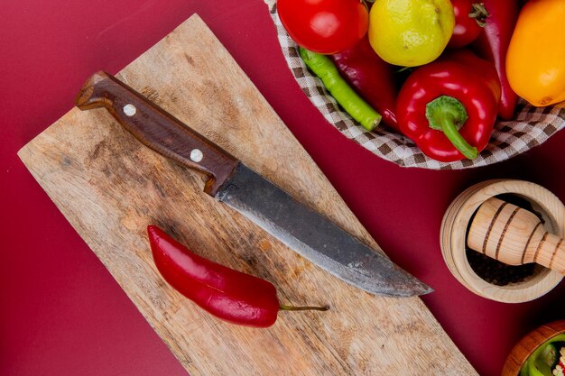 top view of pepper and knife on cutting board with vegetables in basket and garlic crusher on bordo background