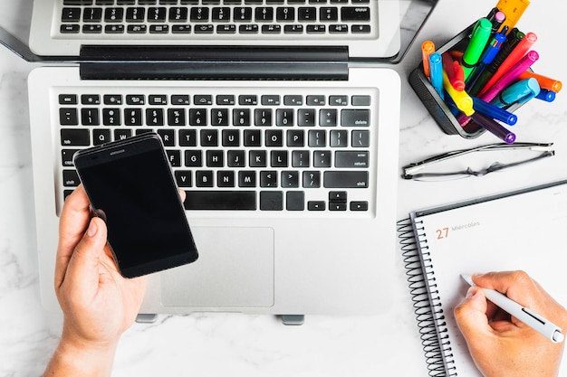 Top view of a person checking his cell phone next to the computer, writing to do in the planner