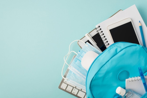 Top view photo of backpack with tablet computer smartphone notebooks pencil pens keyboard mouse sanitizer and medical masks on isolated pastel blue background with copyspace