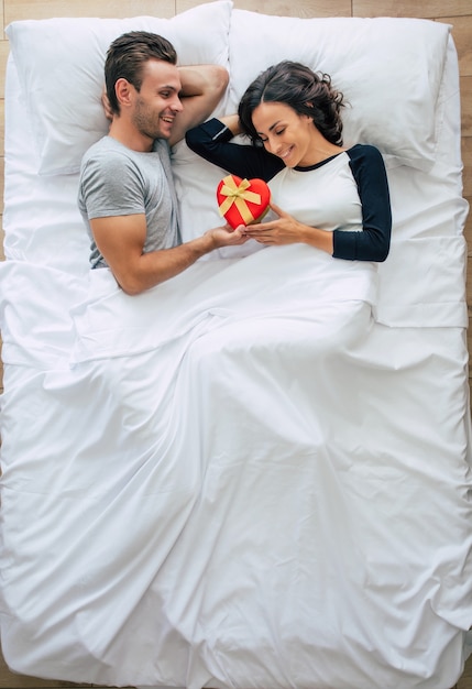 Top view photo of a beautiful happy young couple in love are lying on the big white bed while a handsome man is giving the gift box for his cute woman.