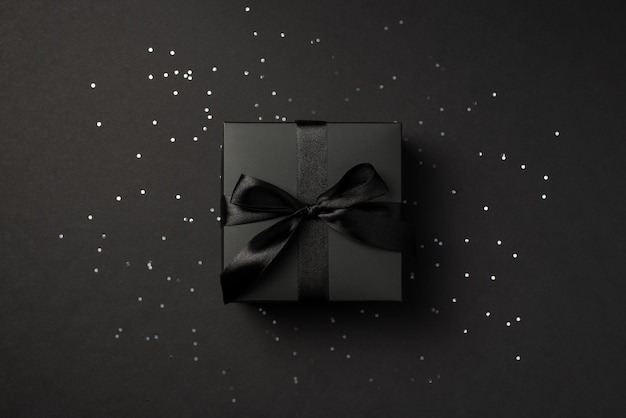 검은색 새틴 리본이 고립된 검은색 배경에 반짝이는 장식 조각 위에 있는 검은색 선물 상자의 위쪽 사진