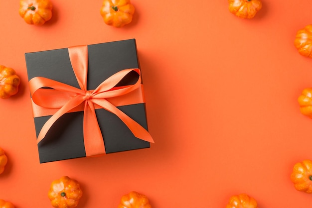 Top view photo of black giftbox with orange ribbon bow and small pumpkins on isolated orange background with empty space