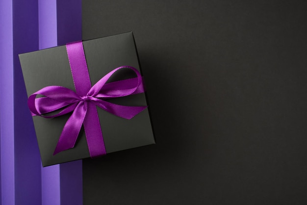 Top view photo of black giftbox with purple ribbon bow on isolated back and violet sheet with vertical folds background with blank space