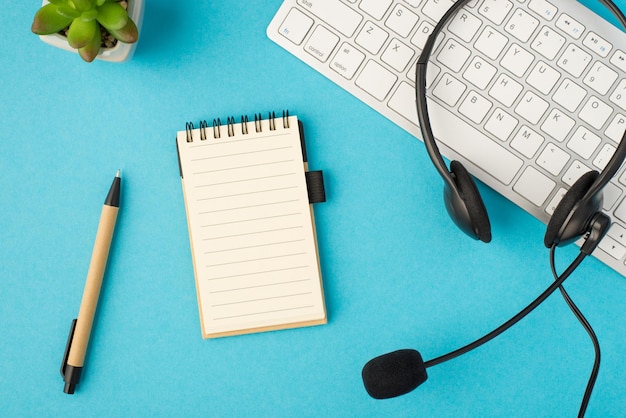 Top view photo of black headset with microphone on white keyboard flowerpot pen and reminder on isolated pastel blue background with blank space