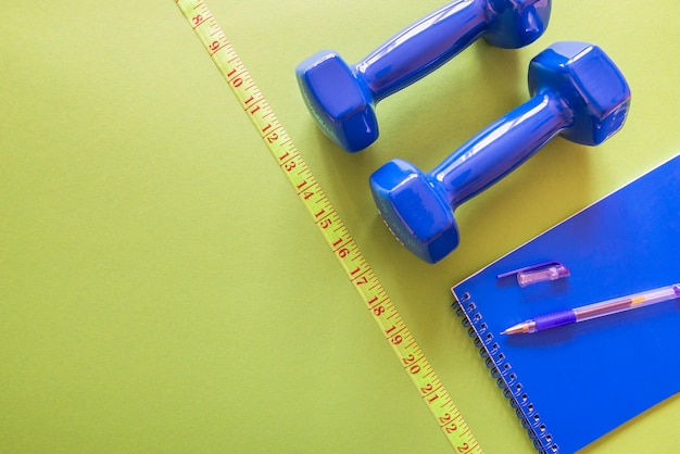 Top view photo of blue dumbbells measuring tape and notepad on a green background with empty spaceFitness and weight loss concept