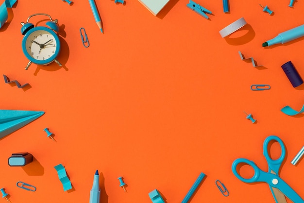 Top view photo of blue school supplies stationery alarm clock markers pencils scissors paper airplane adhesive tape clips and pushpins on isolated orange background with copyspace in the middle