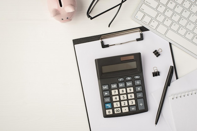 Top view photo of business workplace with keyboard piggy bank glasses pen binders notebook papers and calculator on folder on isolated white background with copyspace