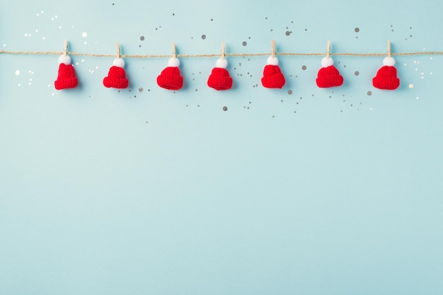 Top view photo of christmas decorations silver confetti and small bobble hats attached to twine with clothespins on isolated pastel blue background with copyspace