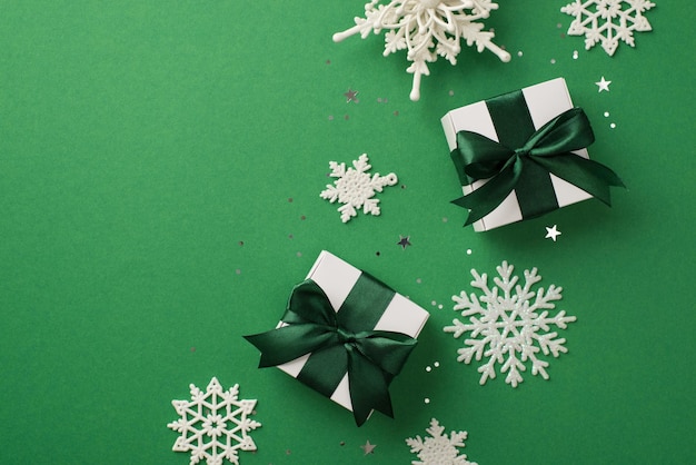 Top view photo of christmas decorations white snowflakes star\
shaped confetti sequins and white gift boxes with green ribbon bows\
on isolated green background with empty space