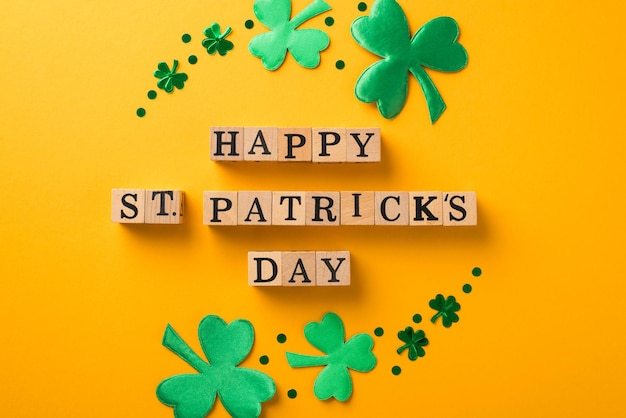 Top view photo of circle composition wooden cubes with words happy st patricks day surrounded by green trefoils and confetti on isolated yellow background